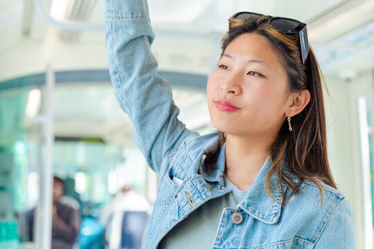 Happy Chinese woman holding onto tram rail with one hand and using smartphone with the other. Smiling in urban public transport. Perfect for lifestyle, technology, travel, and commuting concepts.