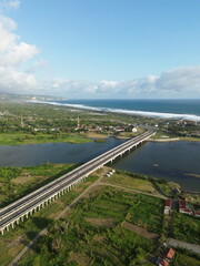 Fototapeta premium Special Region of Yogyakarta, Indonesia - October 15, 2025 : Aerial View Of Coastal Town With Bridge Over Water And Green Fields