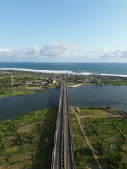 Fototapeta premium Special Region of Yogyakarta, Indonesia - October 15, 2025 : Aerial View Of Coastal Town With Bridge Over Water And Green Fields