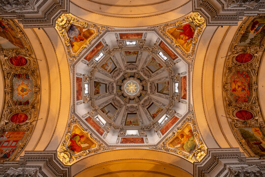 Salzburg, Austria - December 23, 2024: Ornate baroque cathedral ceiling with golden frescoes, painted apostles and intricate stucco decorations illuminated by warm light from high windows above.