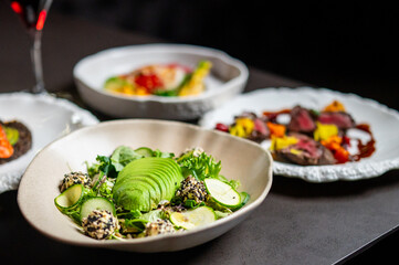 A fresh, healthy avocado salad with greens, cucumber, and seed-covered toppings is shown in a restaurant setting with other gourmet dishes and a glass of wine