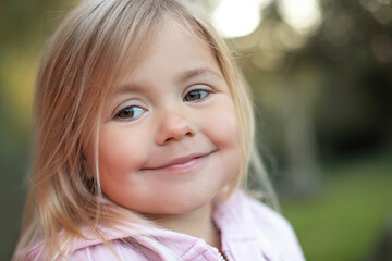 Close-up portrait of a smiling little girl outdoors. Cute child with blond hair and brown eyes looking sideways with a gentle expression. Natural light, soft focus background, warm tones. 