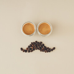 Male face with mustache made of two cups of coffee and coffee beans on beige background. Men's health or father's day concept. Creative Movember awareness idea. Coffee aesthetic. Flat lay. Top view.