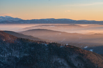 Aerial view of a serene mountain range shrouded in a soft, ethereal mist, where the warm hues of the setting sun kiss the snowy peaks, Lubietova, Banska Bystrica Region, Slovakia.