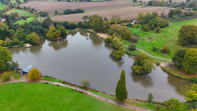Aerial view of a serene lake reflecting the autumnal sky, embraced by emerald green meadows and golden-leaved trees, Pembury Road, Tunbridge Wells, England, United Kingdom.