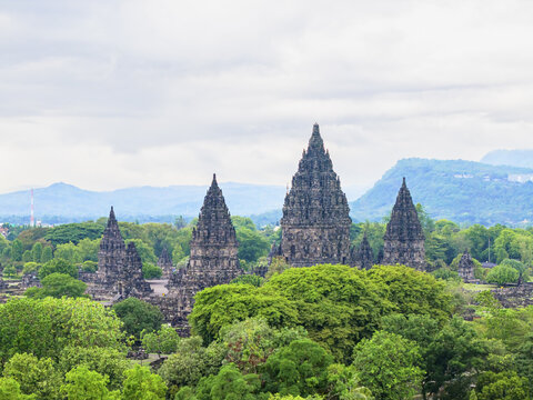Aerial view of the ancient Prambanan Temple's spires piercing through the lush green canopy, set against the backdrop of distant mountains, Yogyakarta, Daerah Istimewa Yogyakarta, Indonesia.