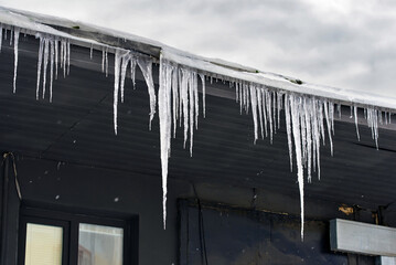 Sharp long icicles hanging from roof edge, serious pedestrian hazard and falling risk as melting ice threatens injury during cold winter weather conditions. Dangerous icicles hanging from rooftop edge