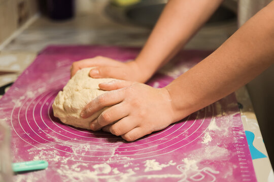 A pair of hands vigorously kneading a mound of fresh dough on a purple silicone baking mat covered in a light dusting of flour.