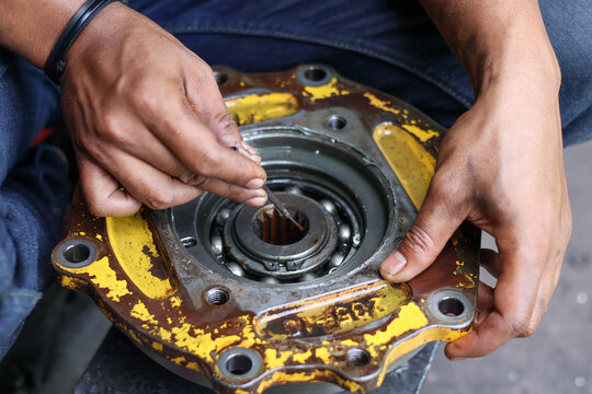 Mechanic's hands using a precision tool to work on a large metal bearing seated within a greasy, worn yellow industrial housing or gear reduction component.