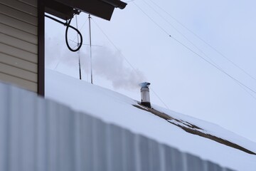 Naklejka premium A chimney on a snow-covered rooftop releases smoke into the cold sky on a winter day.