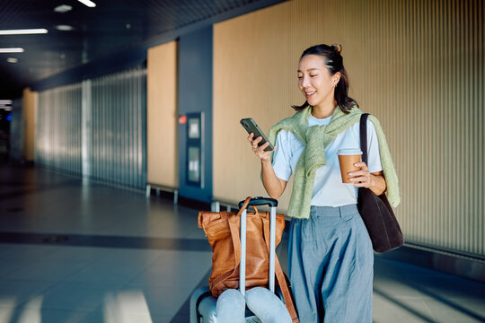 Happy woman passenger standing with luggage and coffee, using smartphone, enjoying travel at a modern airport