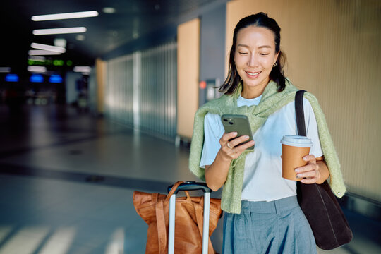 Smiling woman checking smartphone, holding coffee cup, pulling luggage while walking through a modern airport terminal