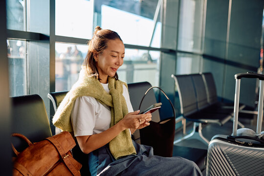 Asian woman traveler relaxing in airport departure lounge, holding passport and smartphone while waiting for boarding