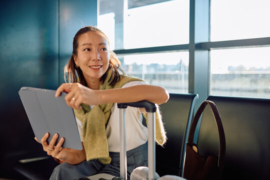 Asian woman traveling, sitting in airport lounge, holding a tablet and smiling while waiting for her flight