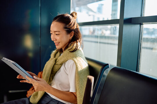 Smiling woman wearing casual clothing sitting in airport lounge, using a tablet computer while waiting for her flight