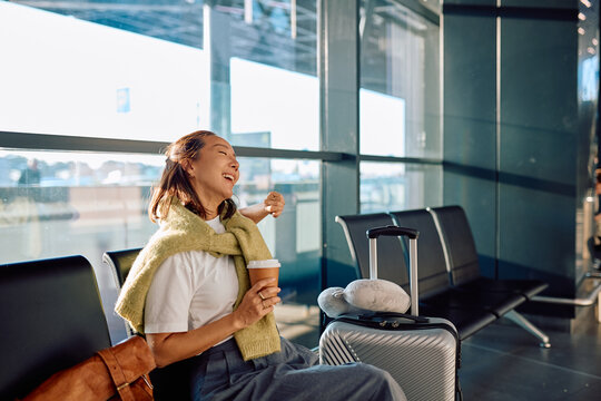 Asian woman laughing while drinking coffee, waiting for her airplane in a modern airport departure lounge with luggage - Powered by Adobe