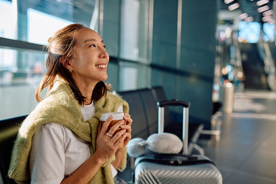 Happy young asian woman passenger resting in airport terminal lounge, holding coffee and waiting for boarding - Powered by Adobe