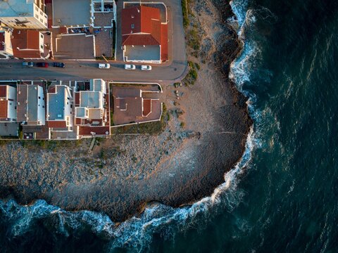 Aerial view of waves crashing against the rugged coastline next to the buildings with red roofs, creating a striking contrast between nature and architecture, Menorca, Islas Baleares, Spain.