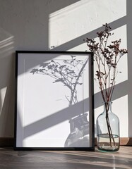 Minimalist black frame mockup with plant shadow and sunlight on a white wall next to a glass vase with dried flowers.