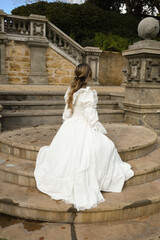 Fototapeta premium back view figure of female blonde model wearing white fantasy wedding gown. romantic sitting pose looking away on staircase of historical fairytale castle location 