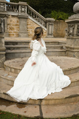 Fototapeta premium back view figure of female blonde model wearing white fantasy wedding gown. romantic sitting pose looking away on staircase of historical fairytale castle location 