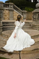 Fototapeta premium back view figure of female blonde model wearing white fantasy wedding gown. romantic sitting pose looking away on staircase of historical fairytale castle location 