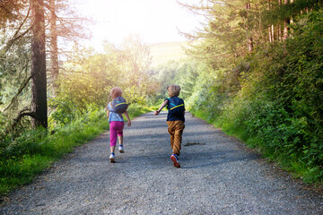 Two kids with backpacks run joyfully along a sunny forest path.