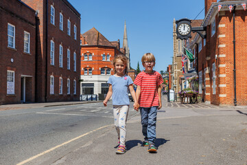 Two kids hold hands, walking down a sunny street of Farnham, UK