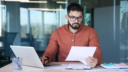 Busy thoughtful businessman analyzing financial documents checking data on laptop while sitting at desk at workplace in a business office. Financier deals with financial reports, engaged in accounting - Powered by Adobe