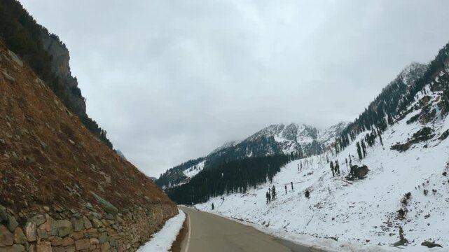 4K POV shot of a vehicle driving through a narrow asphalt road on the side of the Himalayan mountains covered by snow in winter season towards Chandanwari from Pahalgam in Jammu and Kashmir, India.