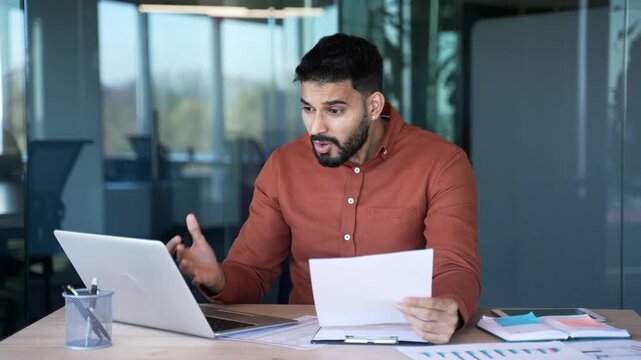 Frustrated businessman unhappy with financial results sitting at desk at workplace in business office. Upset puzzled entrepreneur reviewing documents and looking at laptop screen, unhappy with bad ind