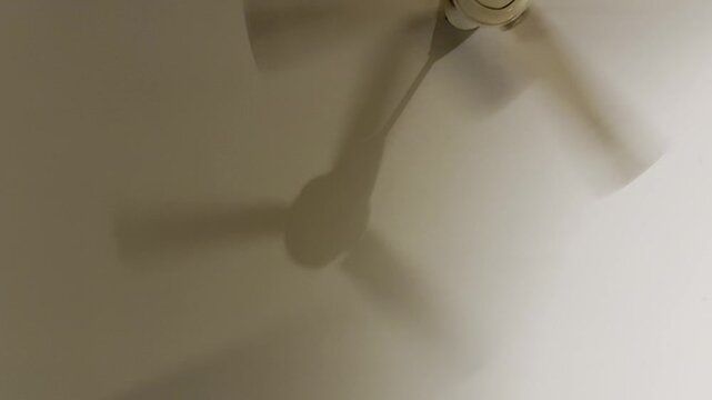 Soft shadow of a ceiling fan slowly rotating on a white wall. Gentle movement creates a quiet, calming atmosphere with warm indoor light.