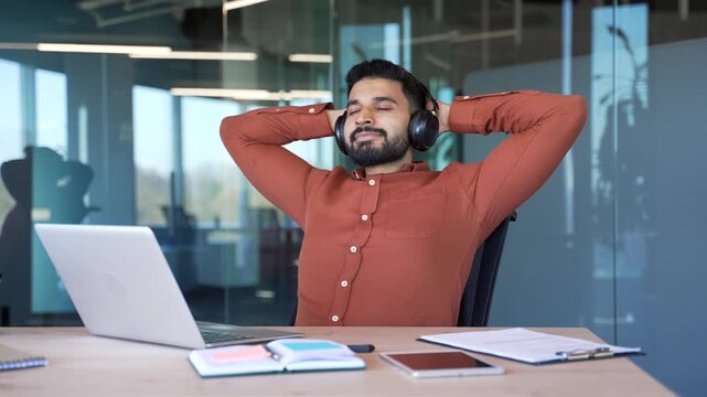 Happy handsome businessman in wireless headphones listening to relaxing music with eyes closed during break at work sitting in office. Satisfied man relaxes to sounds of relaxation and noise of nature