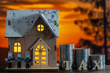 Small toy house model, stack of coins and wooden letters with the inscription tax on table, nature background at night, close up. Concept of land transfer taxation and Investment income tax from asset