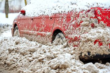 Red car trapped in dirty slush after municipal snowplow clearing operation during heavy winter blizzard, difficult parking and dangerous driving conditions in urban area. Vehicle covered in snowdrift