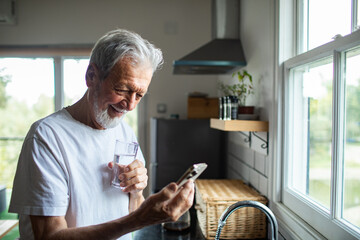 Senior man smiling at smartphone while drinking water in home kitchen