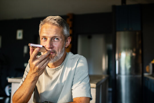 Mature man using voice assistant at home kitchen, thoughtful