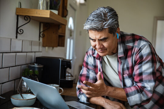 Mature man video calling on laptop in home kitchen, focused