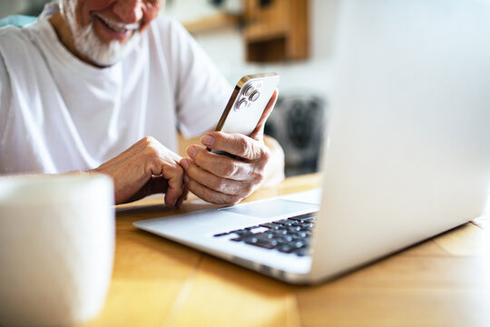 Senior man smiling while using smartphone and laptop at home - Powered by Adobe
