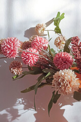 An image of a colorful flower bouquet in the sunlight with pink, mauve, and cream tones. Shadows and light are in the background, on a textured white wall.