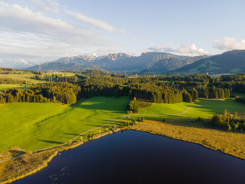Aerial view of a serene lake mirroring the sky, bordered by lush green fields and dense forests, with the majestic Alps rising in the distance, Nesselwang, Bayern, Germany.