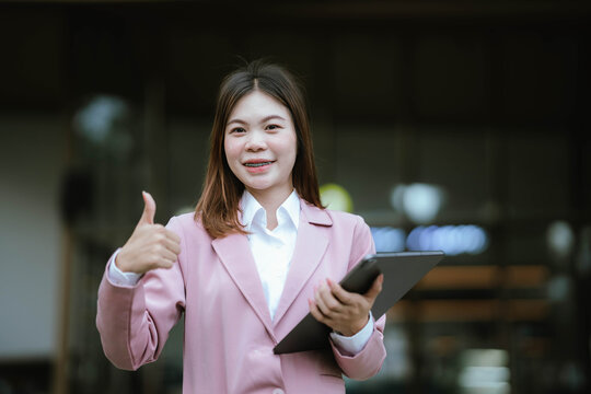 A young woman in business attire smiles while using her smartphone outdoors, holding a pink blazer and appearing engaged and cheerful. - Powered by Adobe