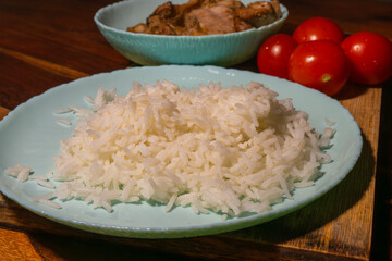 A simple, wholesome meal of rice, grilled chicken, and fresh tomatoes is arranged on a wooden surface with soft natural light.