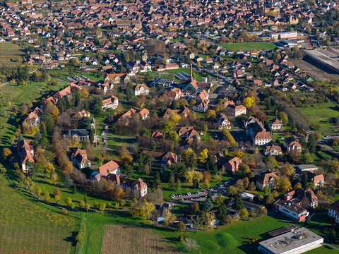 Aerial view of red-roofed buildings nestled in a circular formation amidst lush greenery, a tranquil scene contrasting with the surrounding urban landscape, Rouffach, Grand Est, France.