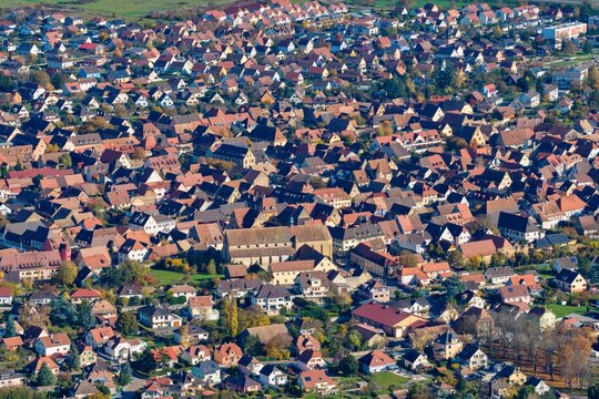 Aerial view of tightly packed buildings and terracotta roofs coalesce around a central church, painting a textured tapestry across the landscape, Rouffach, Grand Est, France.