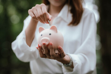 A woman wearing a white shirt is saving money by putting a coin into a pink piggy bank, symbolizing financial planning and saving for the future.