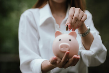 A woman wearing a white shirt is saving money by putting a coin into a pink piggy bank, symbolizing financial planning and saving for the future.