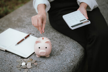 A person saves money by putting coins into a pink piggy bank while holding a calculator, symbolizing financial planning, saving, and budgeting.