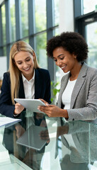 Fototapeta premium Two diverse businesswomen discussing ideas using a digital tablet in a bright office, symbolizing inclusion, leadership, and the global collaboration driving modern professional success.