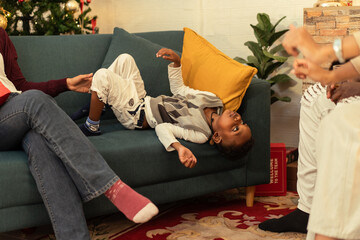 Adorable Black boy relaxing upside down on a couch while spending time with his family. Celebrating...
